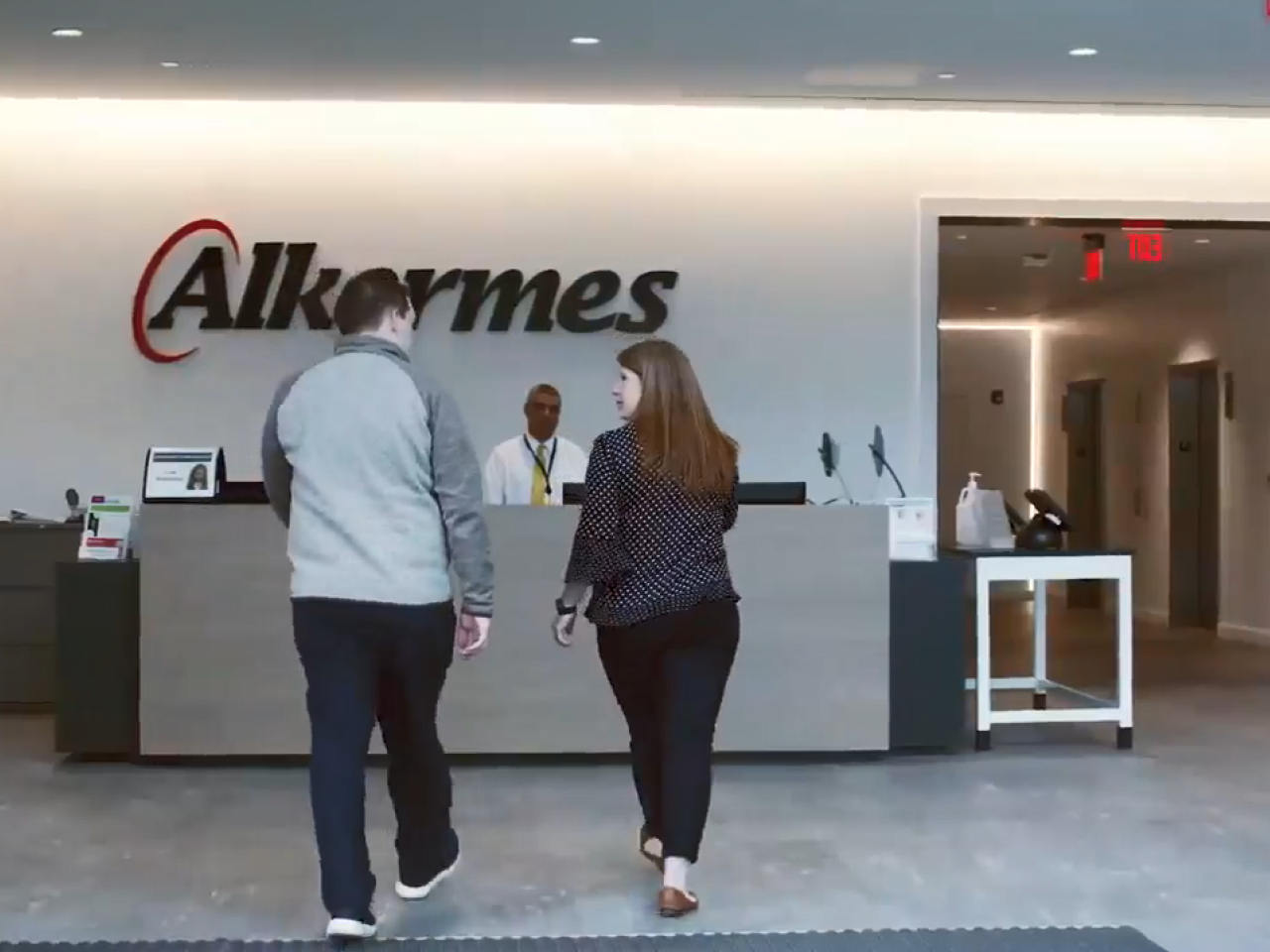 Two people approach a third at a reception desk. A sign "Alkermes" on the wall behind.