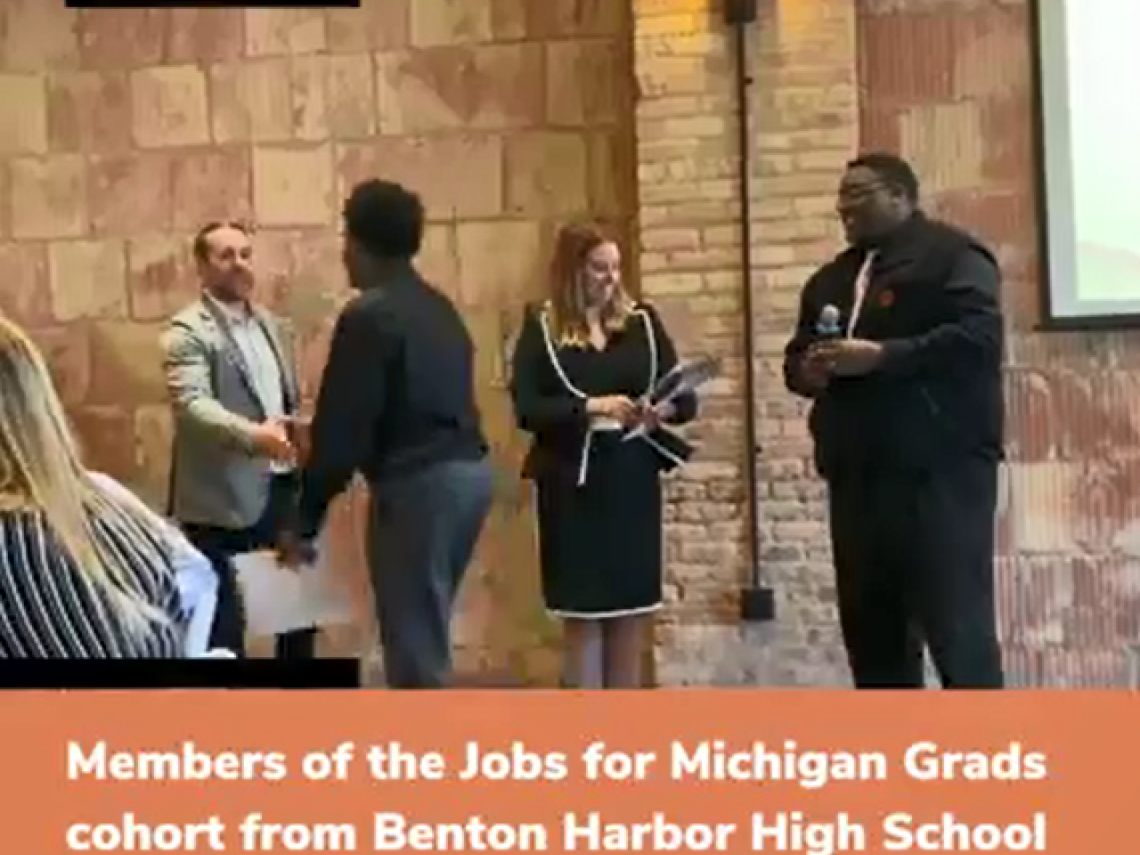 A student walks up to shake hands of three people standing at the front of the room.