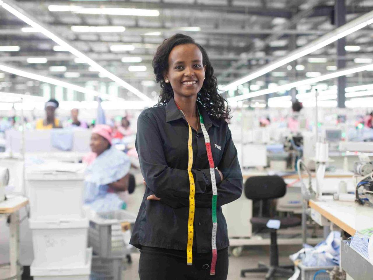 A young women in black clothing stands in front of a large room of women working on sewing machines. 