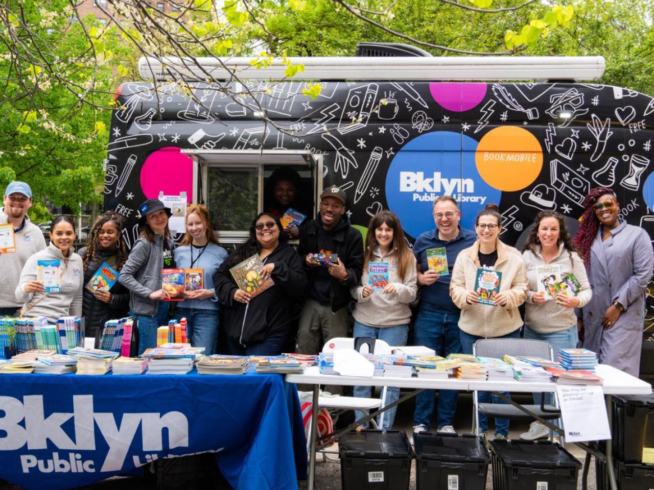 A group of people holding books, stood behind a table that has a Brooklyn Public Library banner draped over 