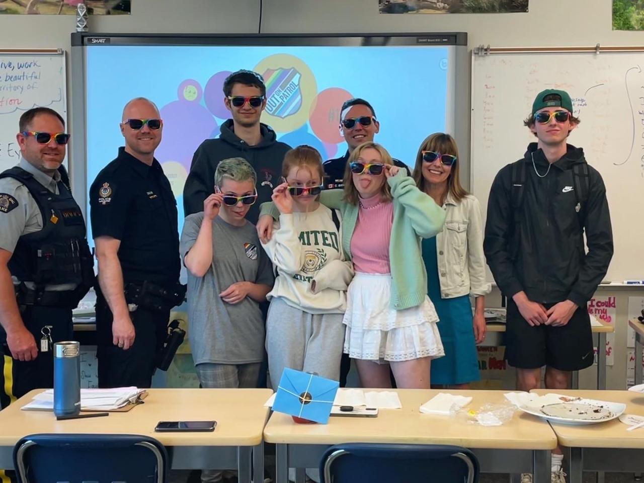 A group of adults and kids all wearing rainbow-rimmed glasses in a classroom setting. Some adults in police uniform.