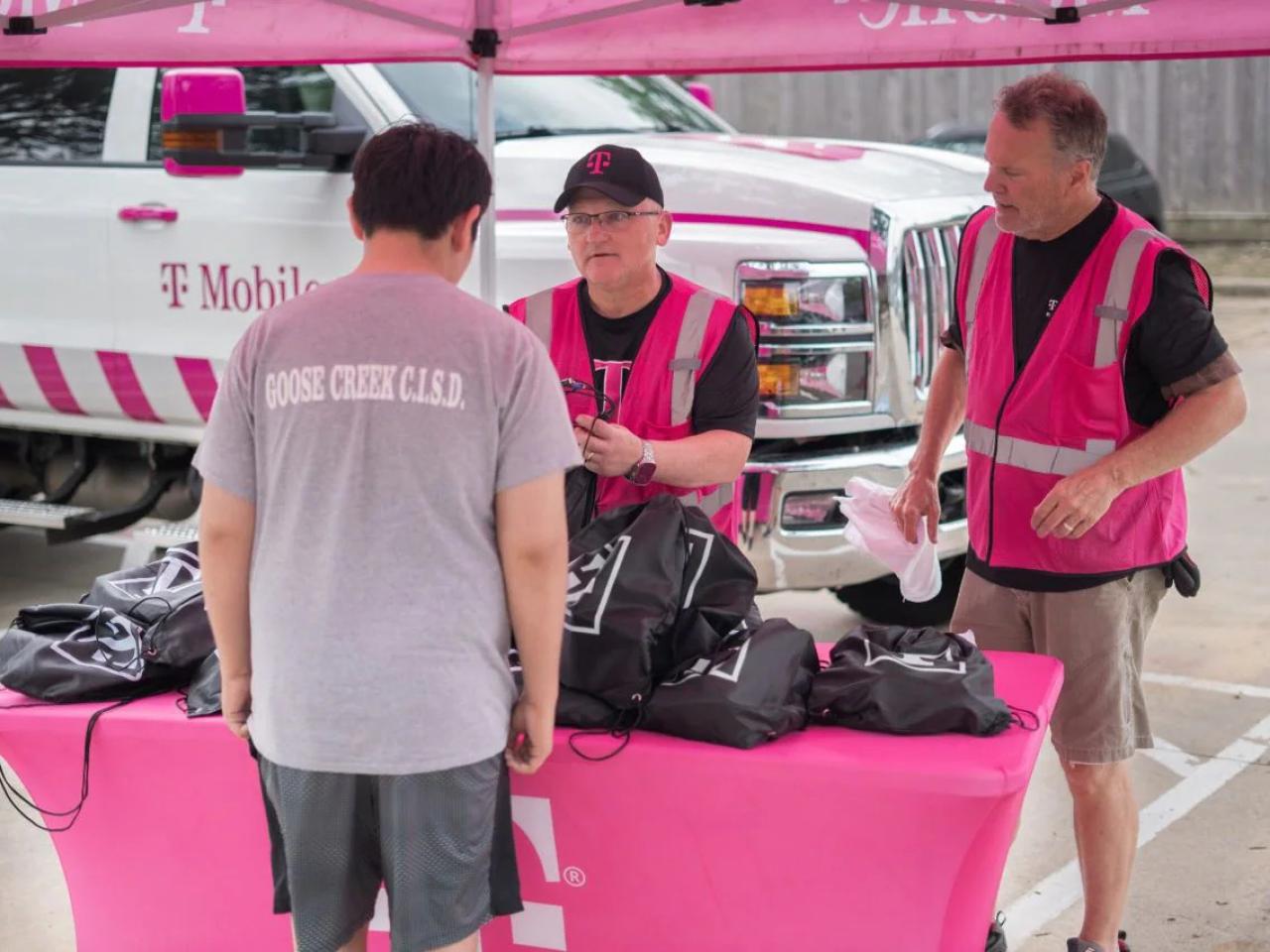 Two people in pink high-vis vests talk to another, a table full of black bags between them. 