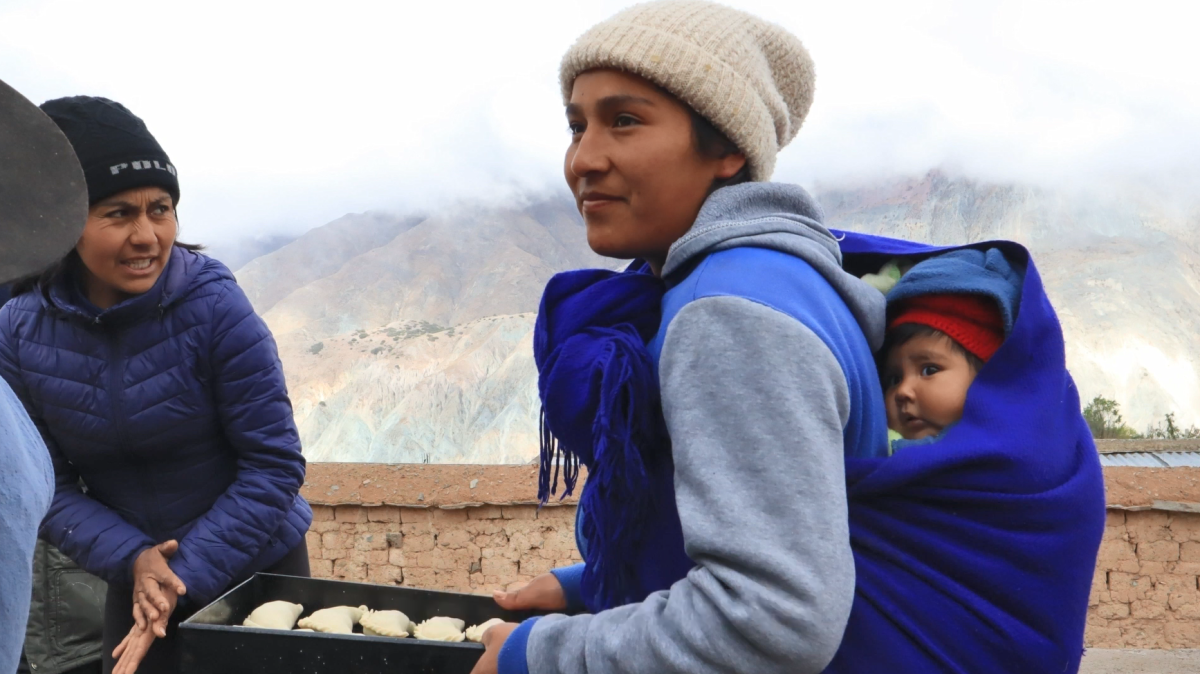 An adult carrying a baby on their pack and a tray of food in their hands, others  greet them.