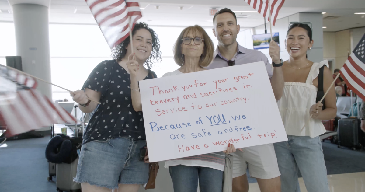 People waving American flags and holding up a sign that reads "Thank you for your great bravery and sacrifices in Service to our country. Because of YOU, We are safe and free. Have a wonder ful trip!"