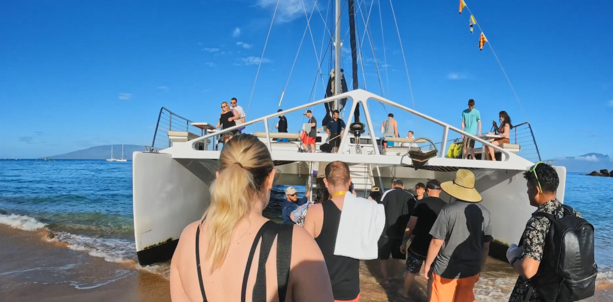 People boarding a large sail boat from a sandy shore.