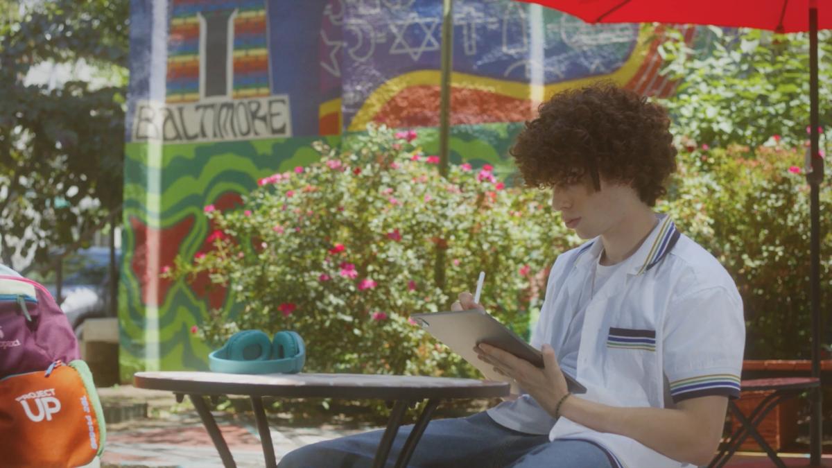 A person seated at a small table with a shade umbrella, using a tablet. A colorful mural on the wall behind them.