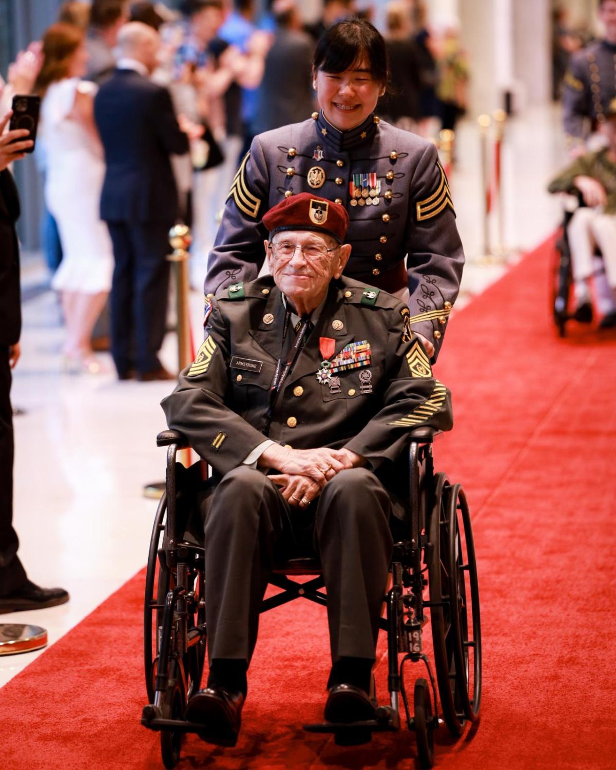 A person pushing a war veteran in a wheelchair down a red carpet 