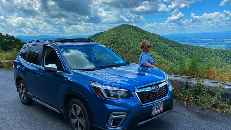 A person leaning against a blue Subaru car, looking out at a long range view