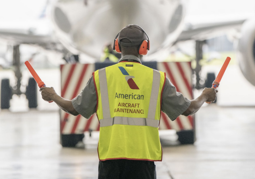 A person guiding a plane wearing a hi-vis vest that reads "American aircraft maintenance" 