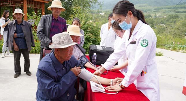 Two elderly people sit at a table outside and have their blood pressure taken by two people in lab coats. A queue of others behind them.