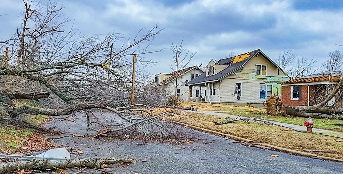 a residential street with three houses all severely damaged by a tornado. A large fallen tree blocks the road.