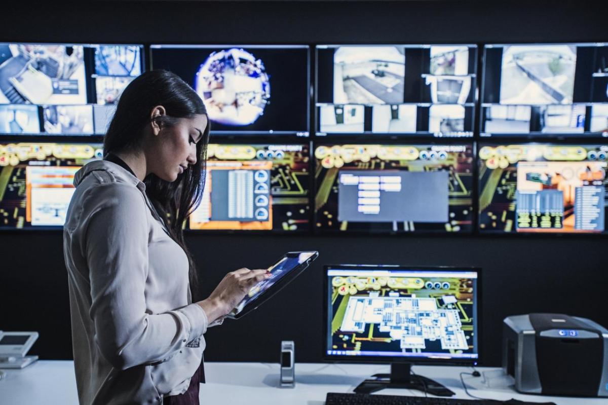 A person using a tablet in a room which has multiple screens on the wall and a computer on the desk