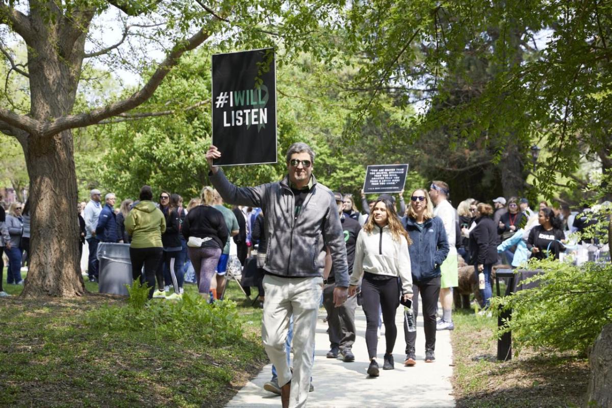 A group walking down a sidewalk and gathered in a park holding a sign "I will listen".