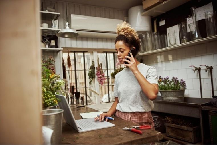 Young woman in a kitchen with her laptop open and speaking on a phone.