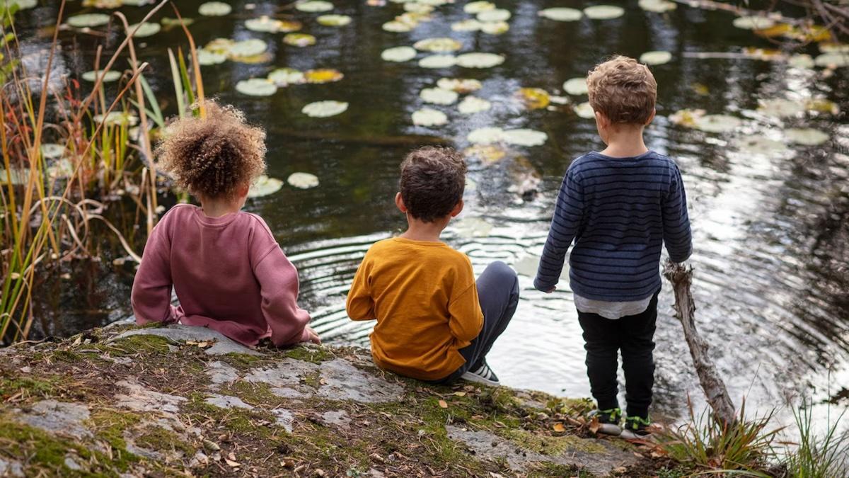 Three small children sitting on the edge of a pond.