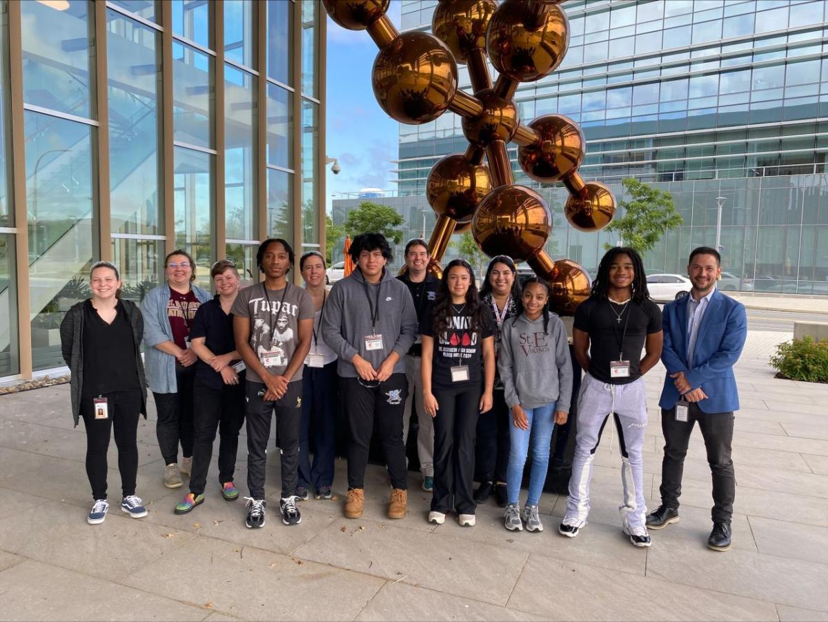 Students and adults posed next to a large molecule statue next to a large glass-sided building.