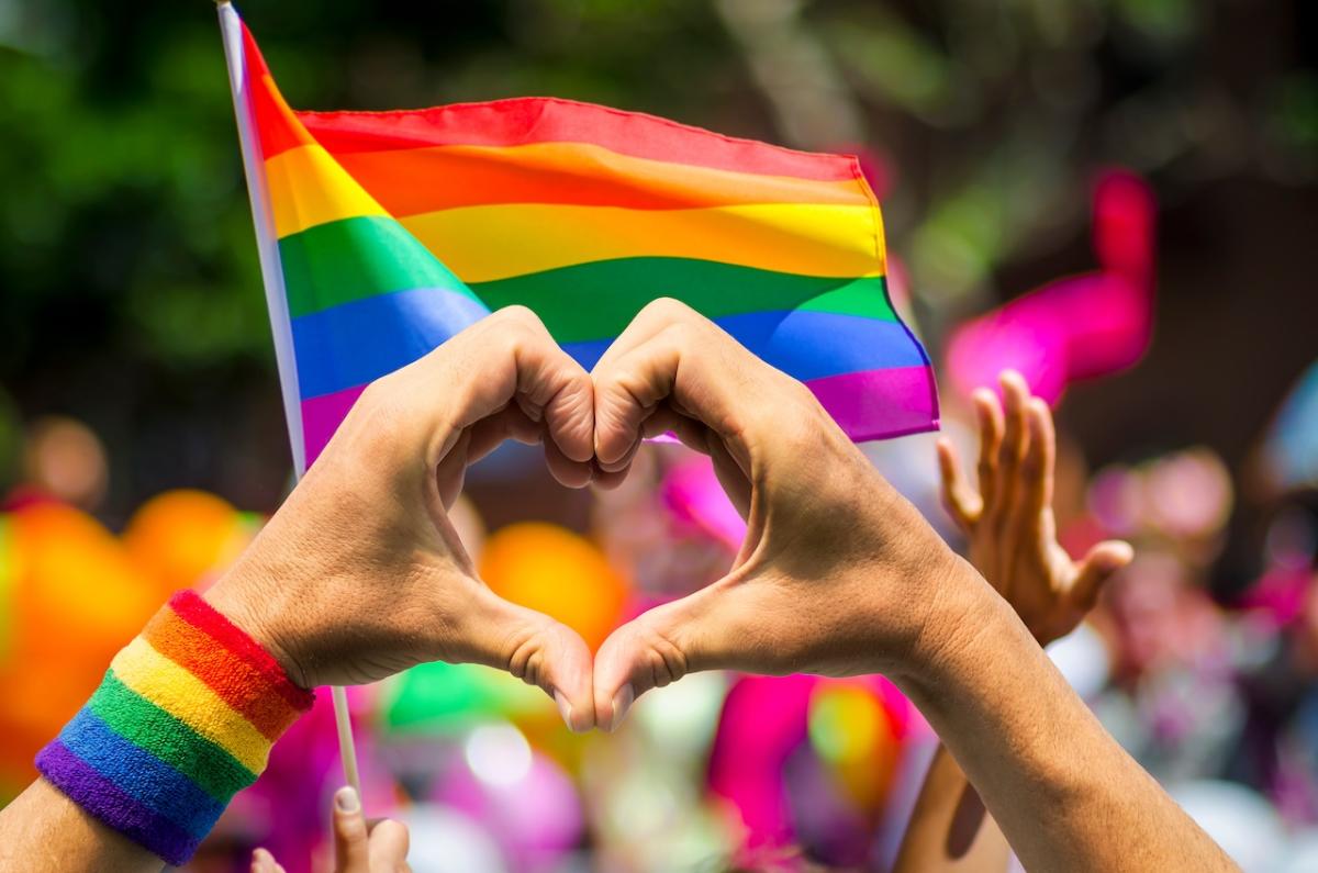 Hand making a heart with a Gay Pride flag in the background.