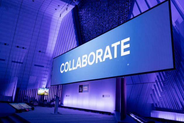  A large digital screen in a modern conference hall displays the word "COLLABORATE" in bold white letters against a blue background. The hall is illuminated with blue and purple lighting, creating a professional and dynamic atmosphere.