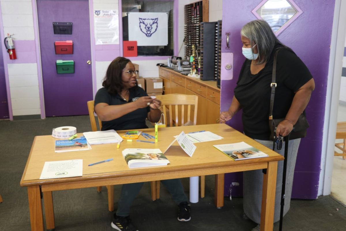 Stacey Ingram sitting at a table with stacks of literature. A person standing beside her wearing a protective mask.