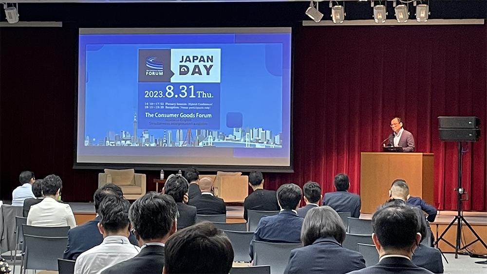 A speaker at a podium in front of a room of seated people. A display to the left showing a presentation.