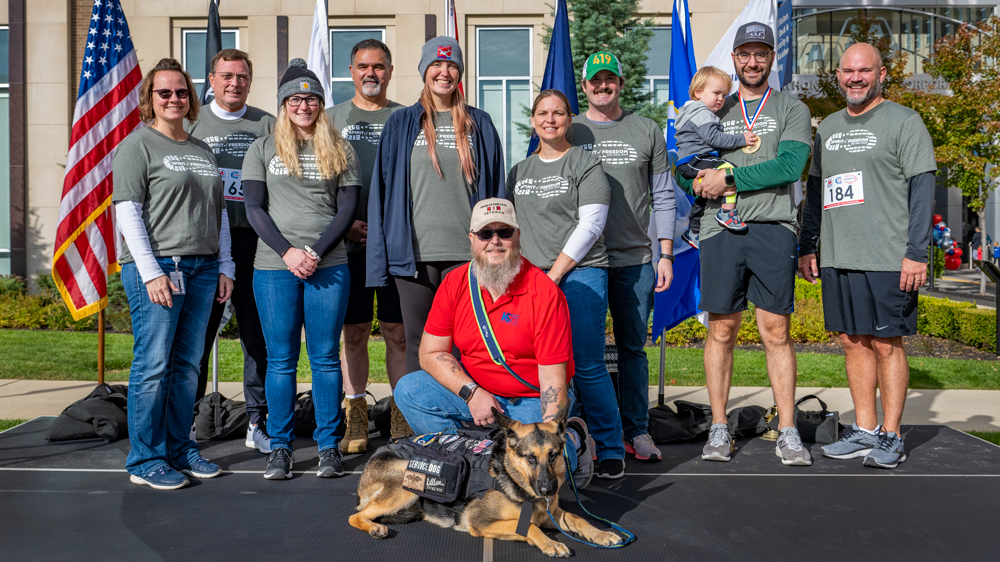 A group of participants, a person kneeling in front of them with a service dog. Flags in the background.