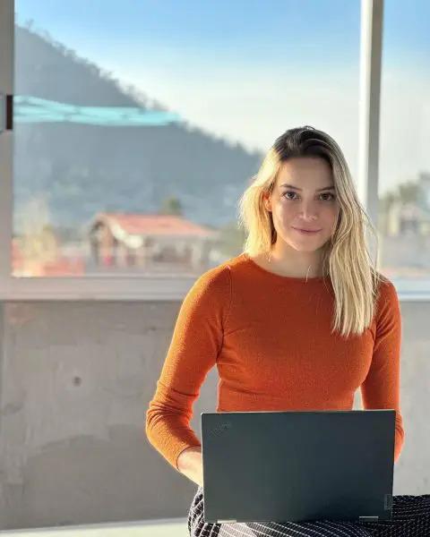 Karla Huerta with an open laptop sitting on a bench in front of a large window.