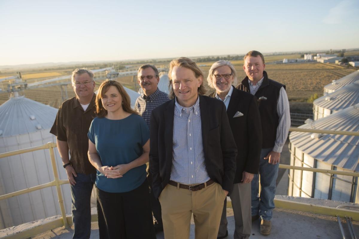 Group of six people stand on a rooftop overlooking storage silos and a field