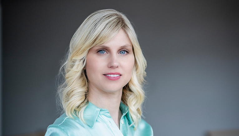 Martina wearing a shiny pale green shirt, a grey background behind her.