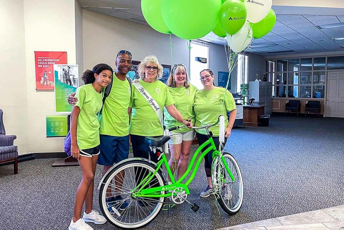 Mary Lou Schlueter and family pose in green t-shirts behind a green bike and balloons