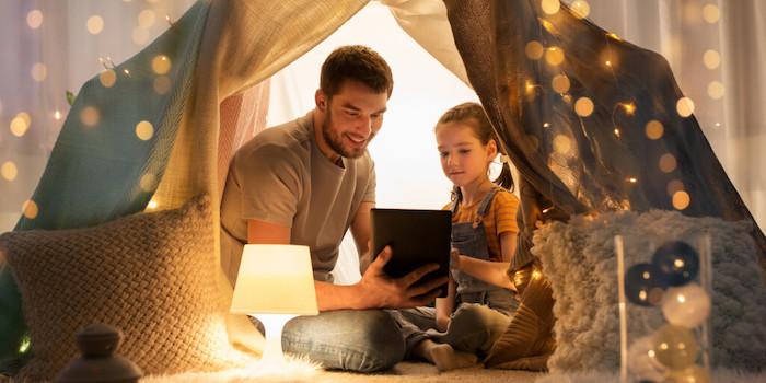 Father and daughter seated behind a curtain and looking at the screen of a laptop.