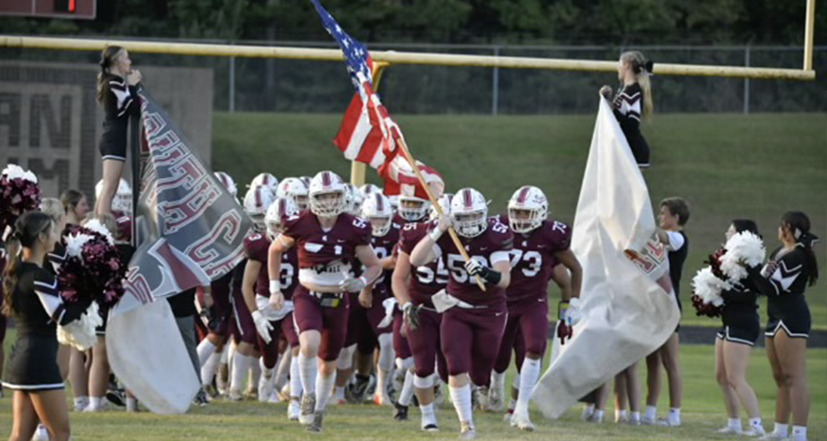 A football team running onto a field, one carrying an american flag.