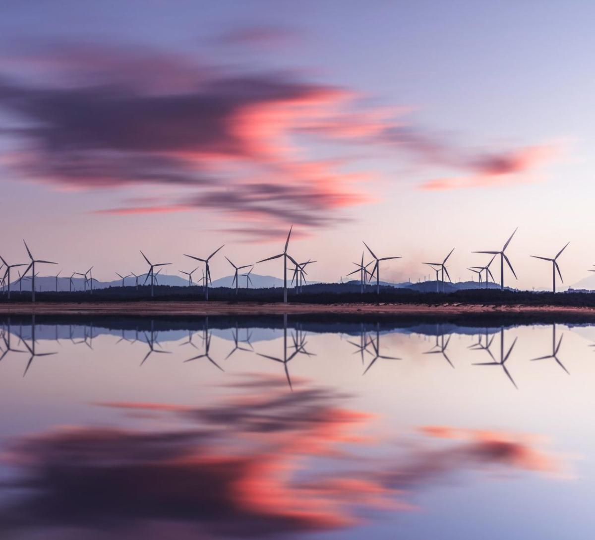 Wind turbines behind a body of water. A setting sun behind them.