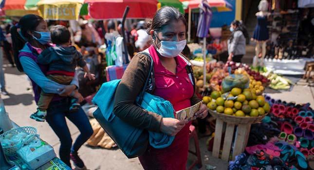 Person walking through a food market carrying a bag. A person behind them carries a young child.