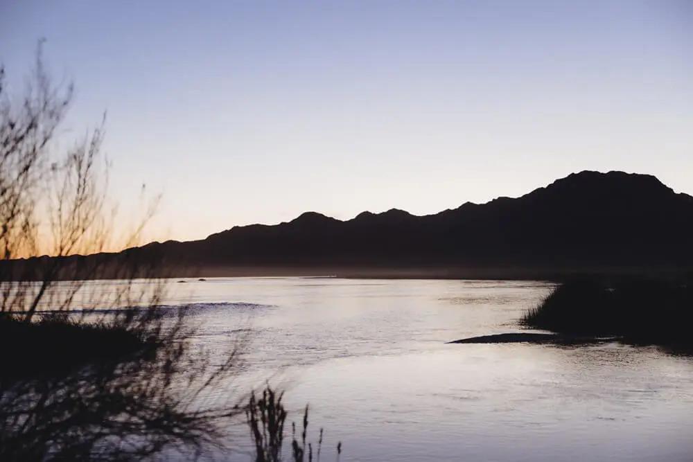 Landscape of a waterway, tall grass, and hilly terrain. A setting sun to the side.