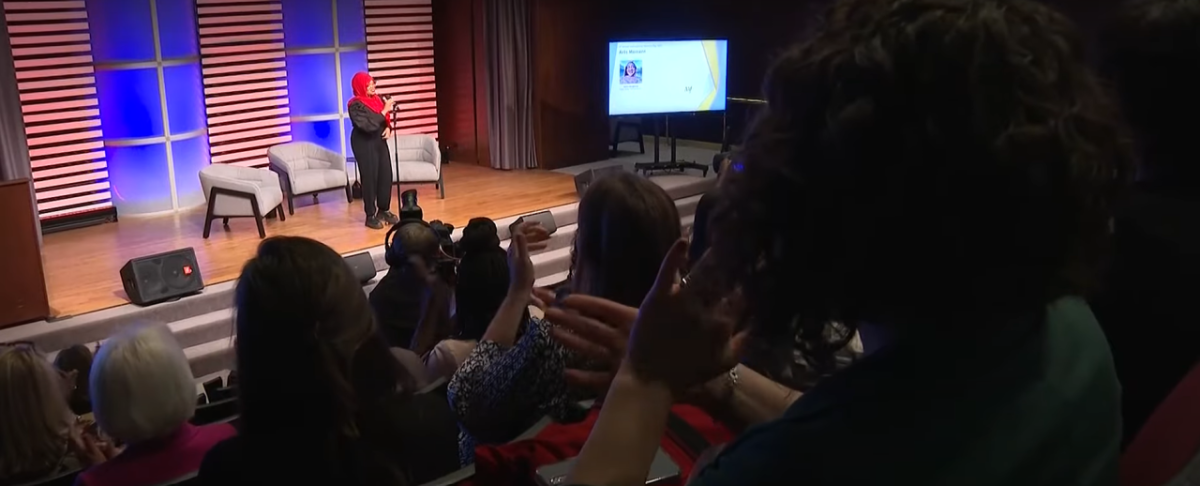 Saleemah Abdul-Ghafur speaking on a stage with three armchairs. An audience in front of her.