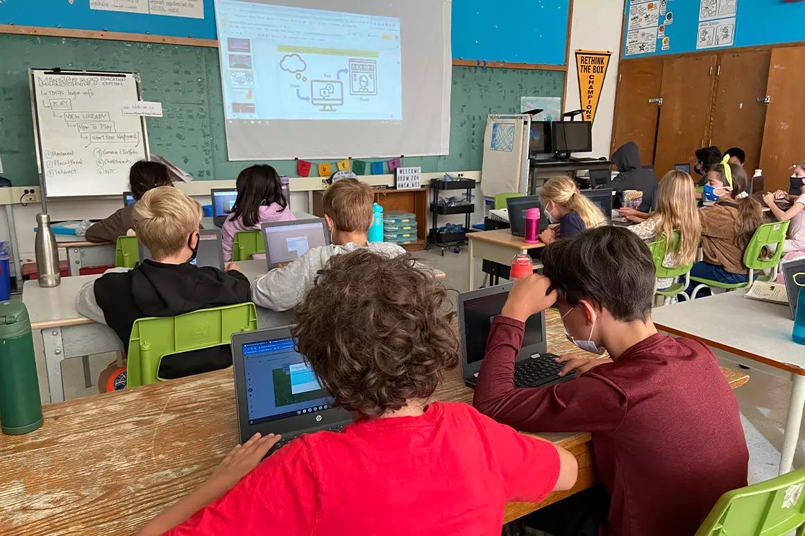 a classroom of students sitting in pairs, each on an electronic device. A lesson projected on the screen at the front of the room.