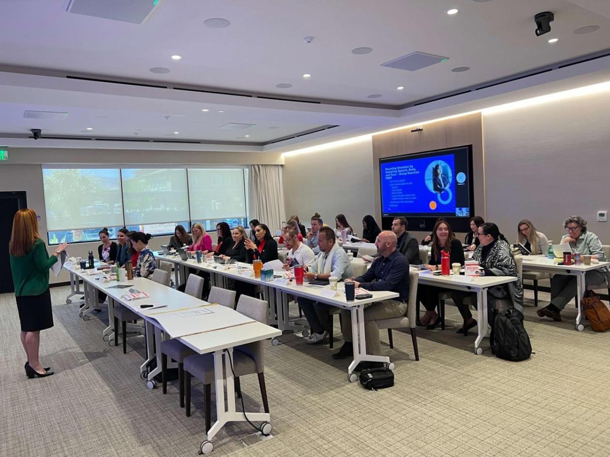 A group seated at tables in a conference room. One speaking in front.