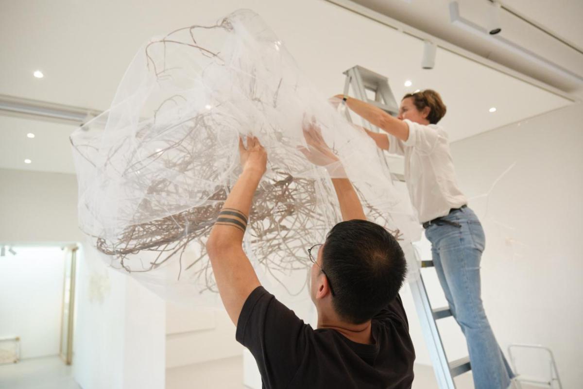 two people attach a gauzy textile sculpture to a gallery ceiling