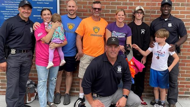 A group of adults and children smiling at the camera. Some wearing police badges. A brick building behind them.