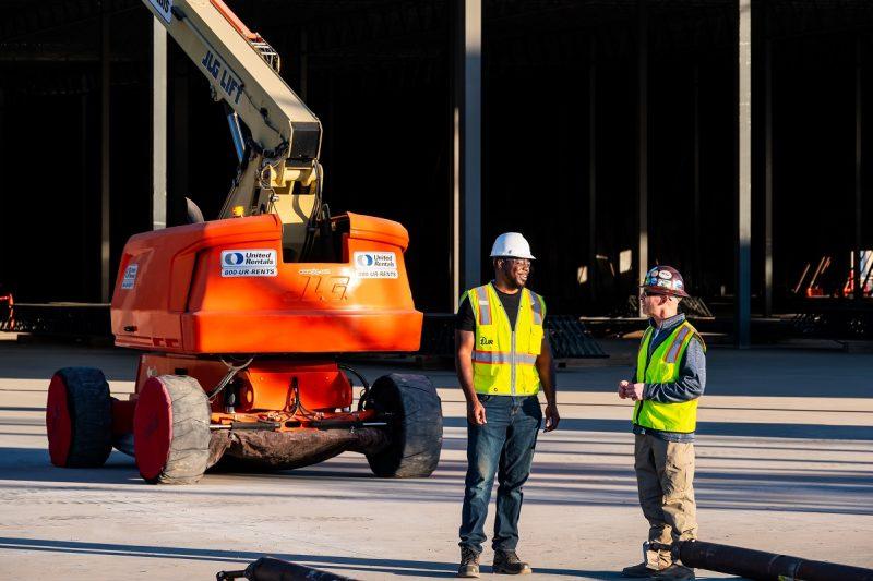 Two workers in hard hats and high vis vests stand in front of a large lift