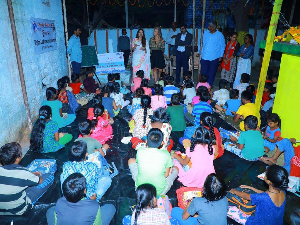 A group pf students, seated, adults standing at the front of the room.