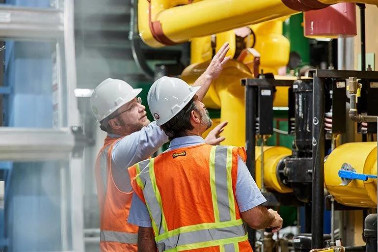 Two people looking at large yellow indoor piping, each wearing hard hats and high-vis vests.