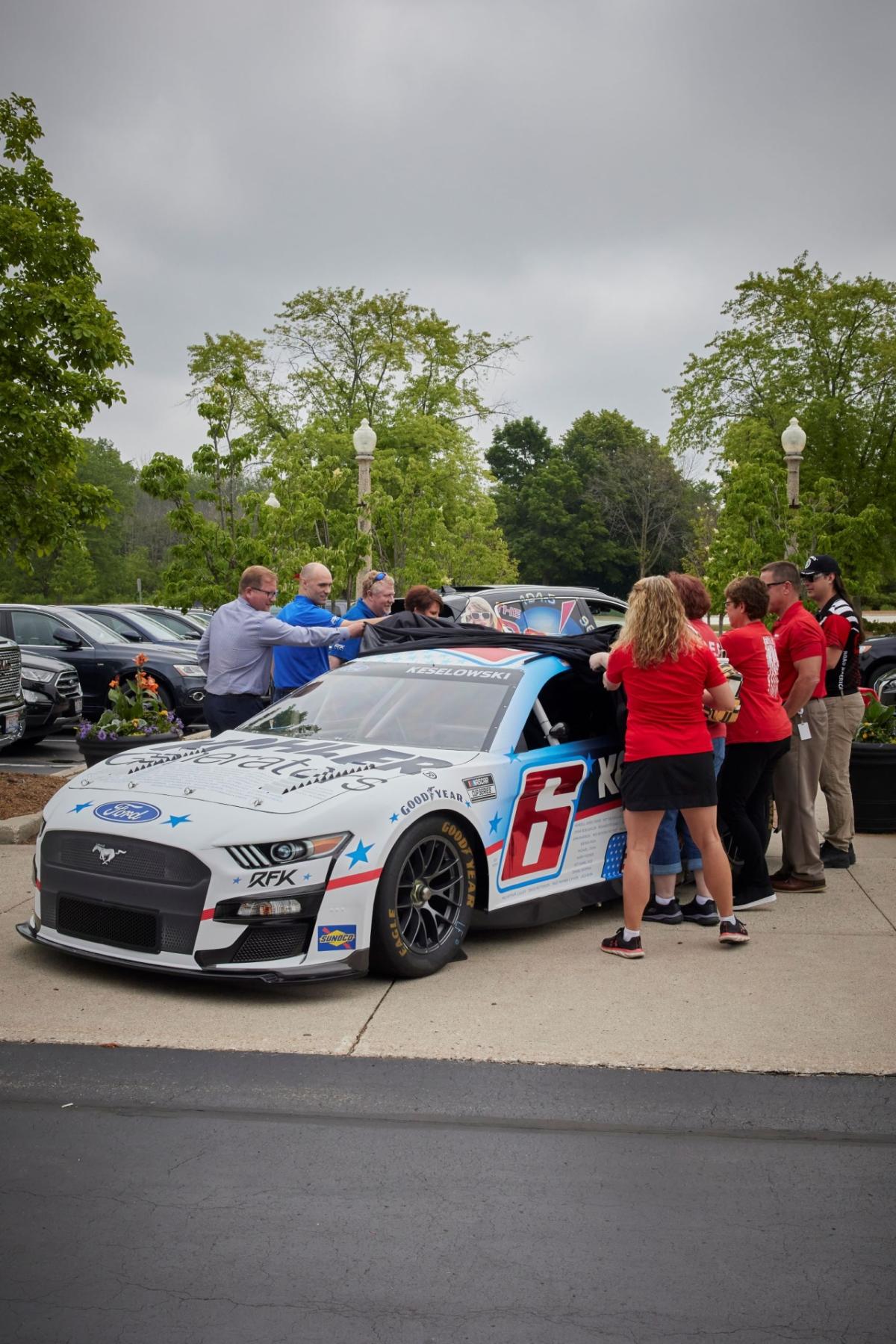 a group of people stand outside and take a cover off a race car. "6" on the side