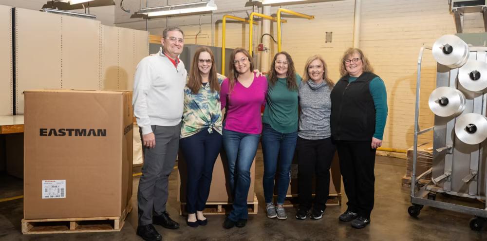 Six people posed with arms around each other. Large spools of thread to the side and an Eastman Box on the other side.