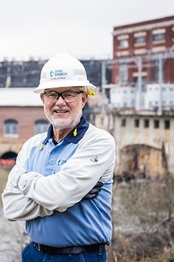 A smiling person with arms crossed in a hard hat. A hydroelectric plant behind them.