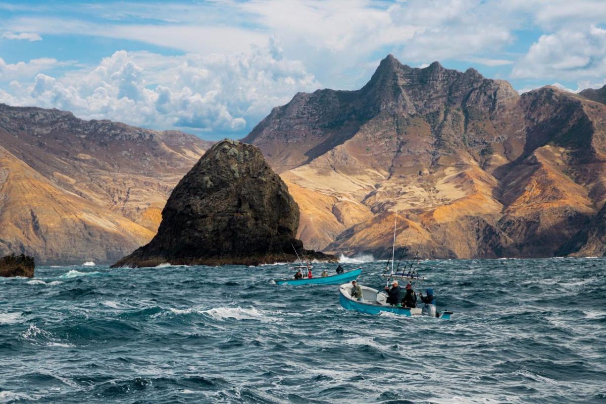 Two small boats carrying people on wavy water. Large mountains in the distance.