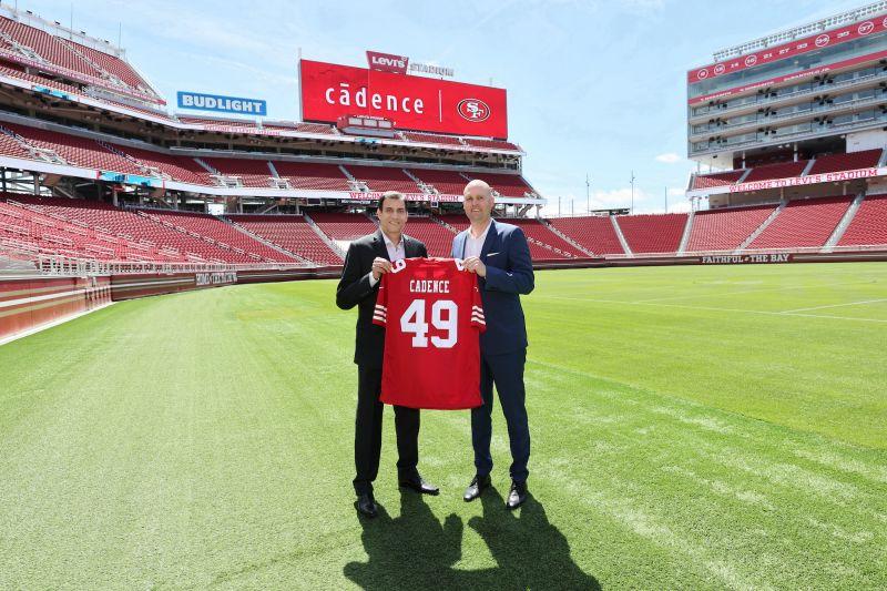 Two people holding a red jersey with 49 on it, standing on a football field. Large "cadence" billboard at the top of the stands.