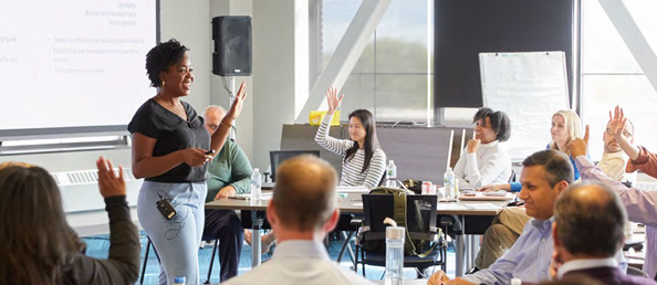 A person with hand raised speaking to a room of adults, some with their hands raised.