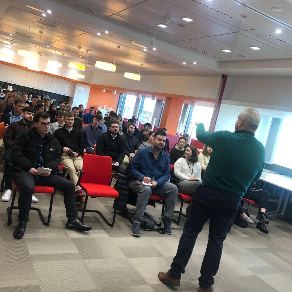 A room full of seated students and staff. A person with a microphone speaking in front of them. 