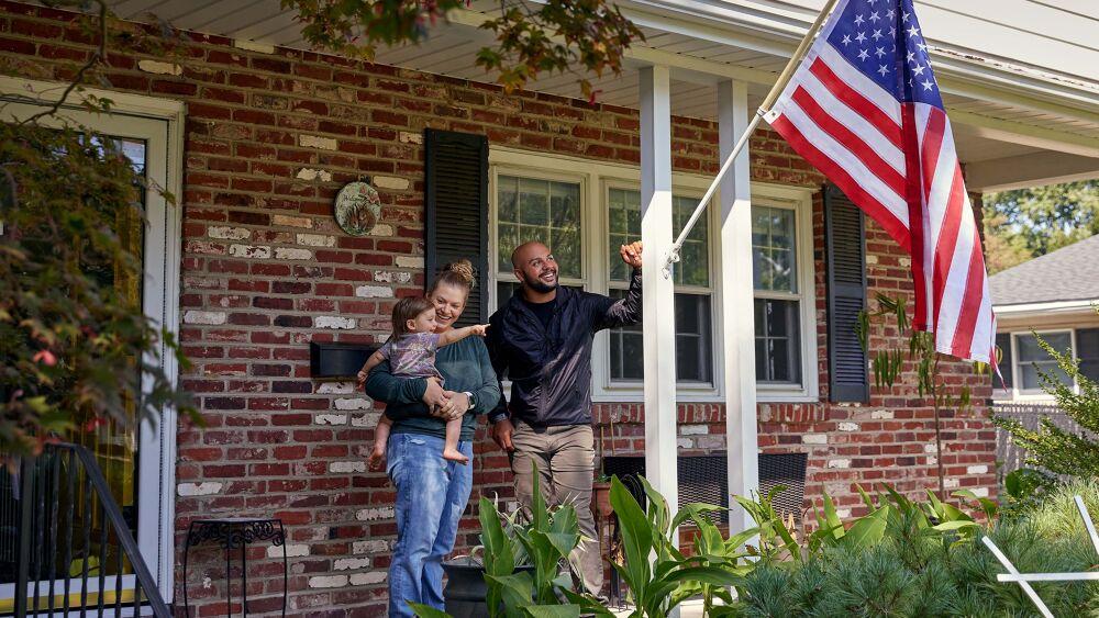 Two adults and a small child standing on a porch. An american flag in a post on the side pillar.